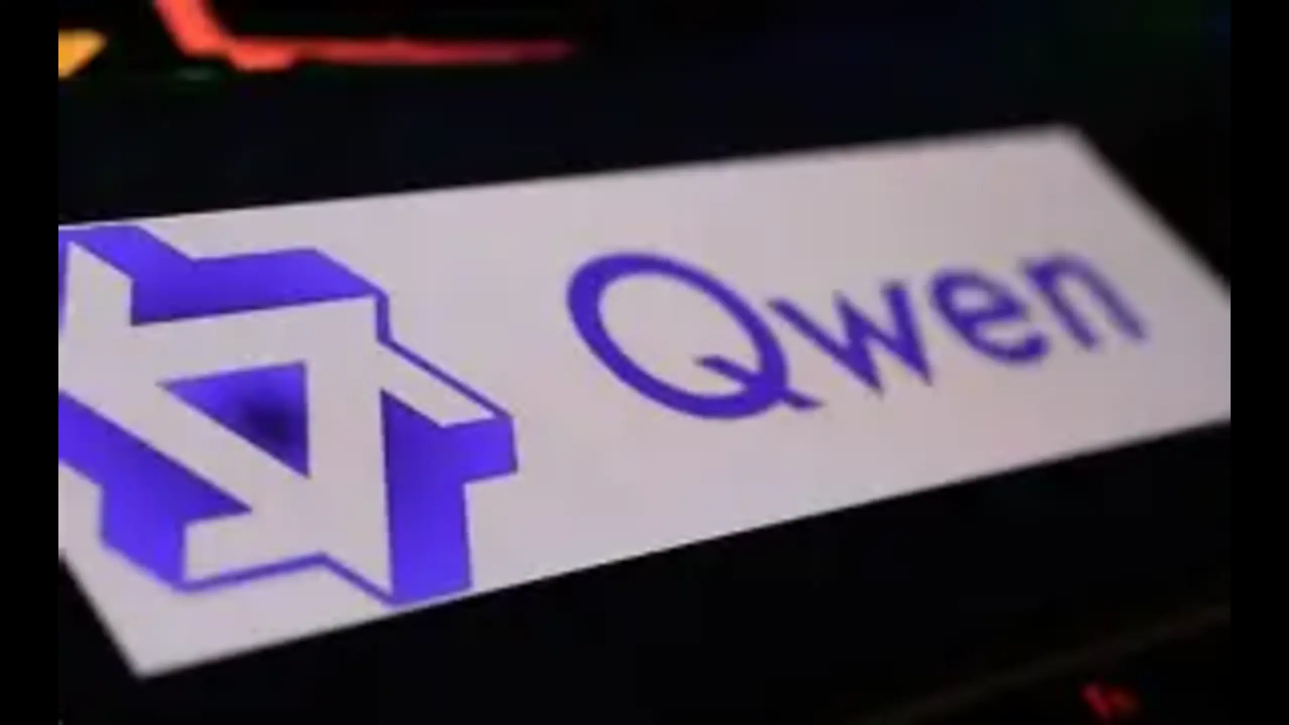 A close-up of a sleek nameplate featuring the word "Qwen" in bold purple text alongside a modern, geometric blue logo design, set against a dark background, representing the Qwen3 coder AI model.